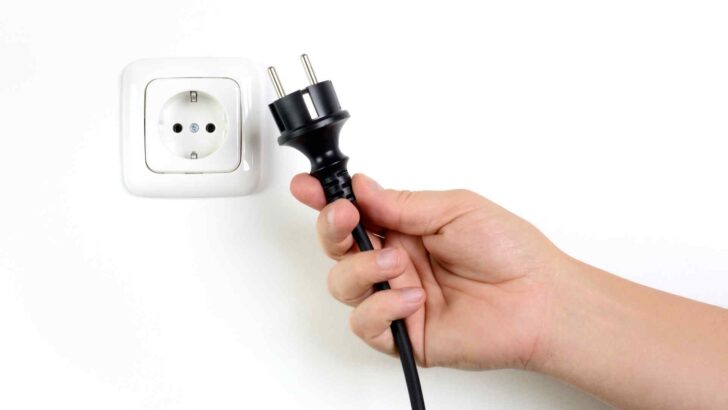 A hand holding an unplugged power cord beside a wall outlet