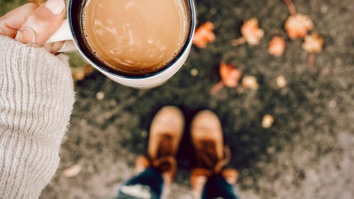 A person holding a warm drink above fallen autumn leaves